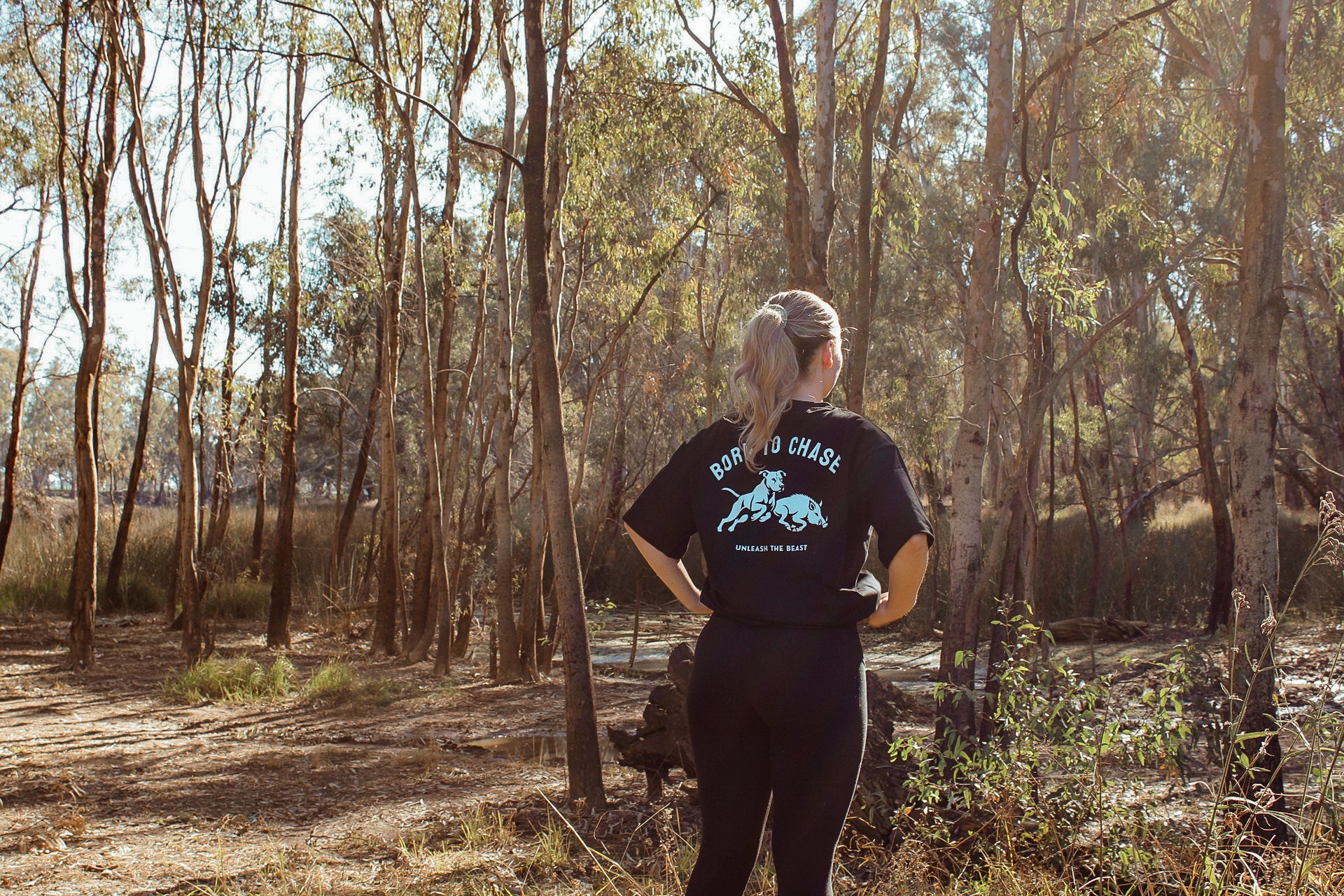 Woman wearing the black Born to Chase T-shirt from The Boar Collective standing in natural Australian bushland.