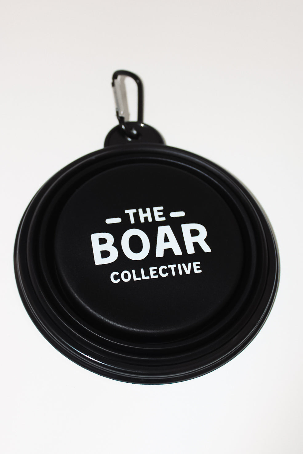 Front view of folded matte black collapsible dog bowl with white The Boar Collective logo and silver carabiner
