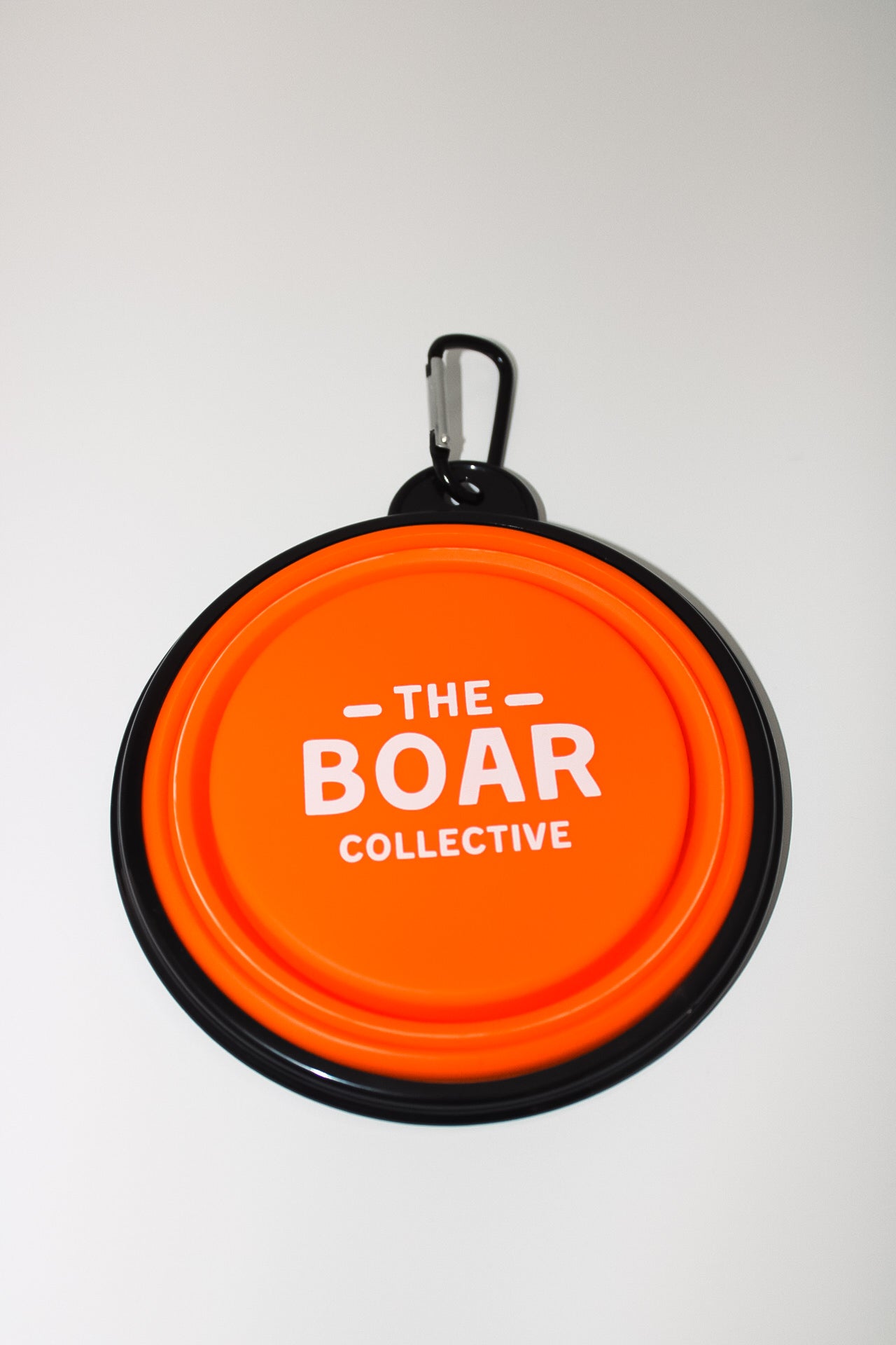 Folded blaze orange collapsible dog bowl with white The Boar Collective logo and carabiner clip, top-down view